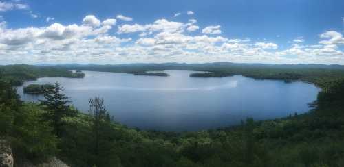 Panoramic view of a serene lake surrounded by lush green forests and distant mountains under a blue sky with clouds.