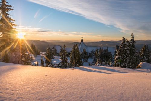 A snowy landscape at sunrise, with a church nestled among evergreen trees and mountains in the background.