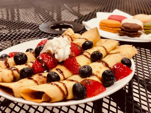 A plate of crepes topped with strawberries, blueberries, whipped cream, and chocolate sauce, with macarons in the background.
