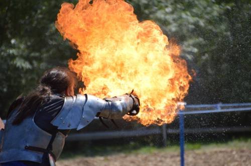 A person in armor shoots a burst of fire from their hand, surrounded by greenery and a blurred background.