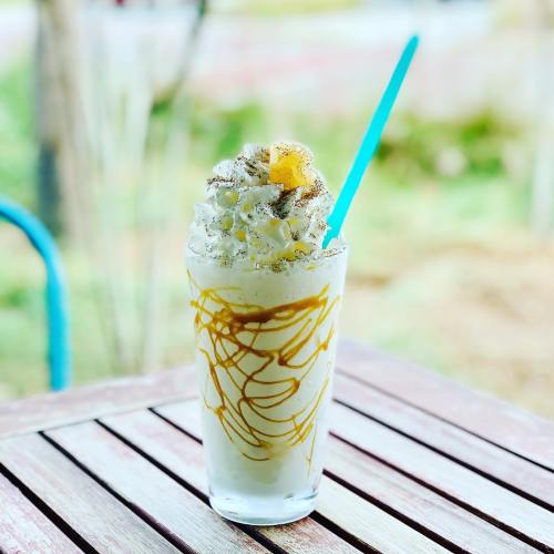 A tall glass of creamy dessert topped with whipped cream and drizzled with caramel, served with a blue straw.