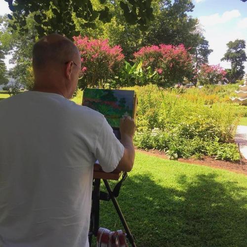 A person stands outdoors, painting on a tablet in a lush garden with colorful flowers and greenery.