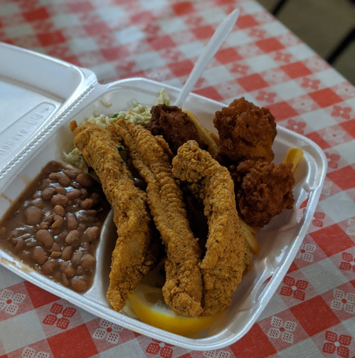 A takeout container with fried fish, hushpuppies, coleslaw, and baked beans on a checkered tablecloth.