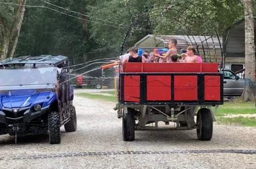A blue ATV and a red trailer filled with people spraying water at each other on a gravel road in a wooded area.