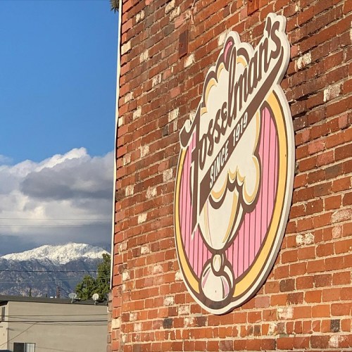 A vintage ice cream shop sign on a brick wall, with snow-capped mountains in the background under a clear sky.