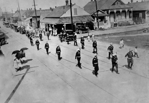 Historic black-and-white photo of a parade with marching soldiers, vintage cars, and spectators on a street.