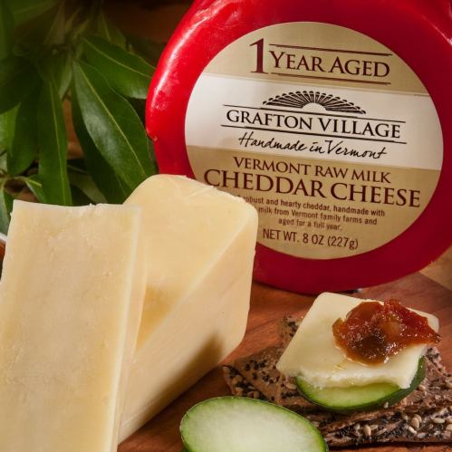 A round wheel of aged cheddar cheese with a wedge cut out, accompanied by a cucumber slice and chutney on a wooden surface.