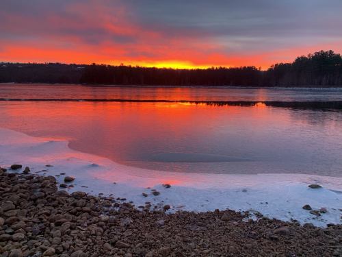 A serene sunset over a calm lake, with vibrant orange and purple hues reflecting on the water and a rocky shore.