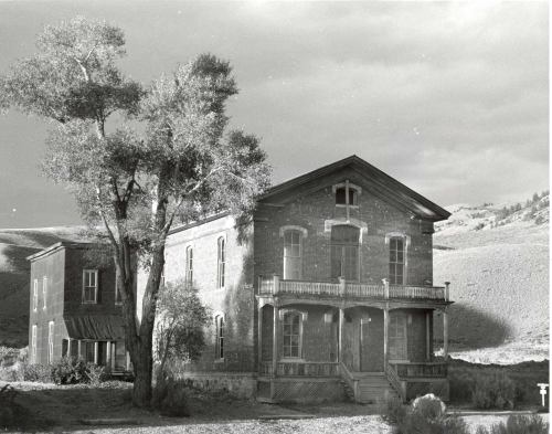 A historic two-story brick building with a porch, surrounded by trees and hills, under a cloudy sky.