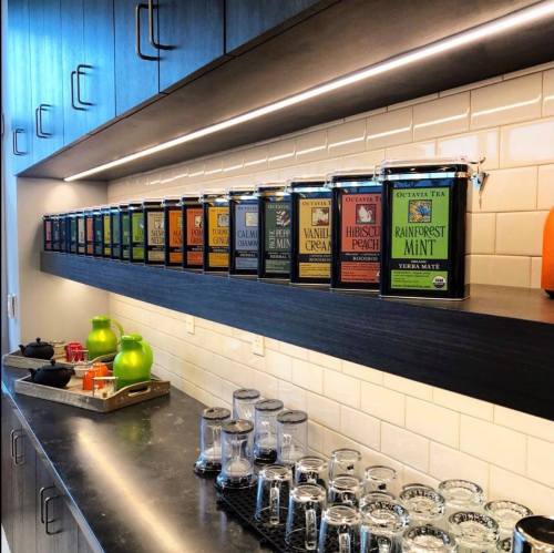 A modern kitchen shelf displaying various tea canisters in colorful packaging, with glass jars and green apples below.