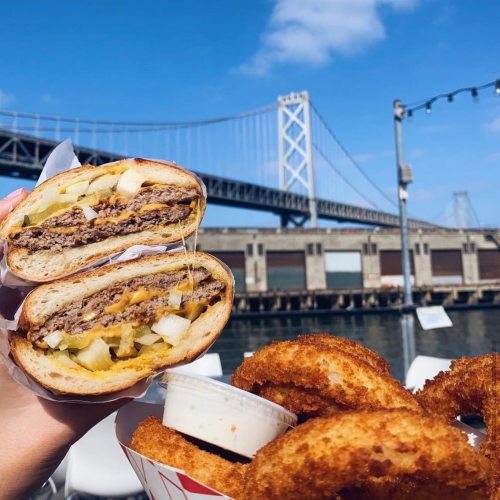 A hand holding a cheeseburger with pickles and onions, alongside a serving of crispy onion rings, with a bridge in the background.