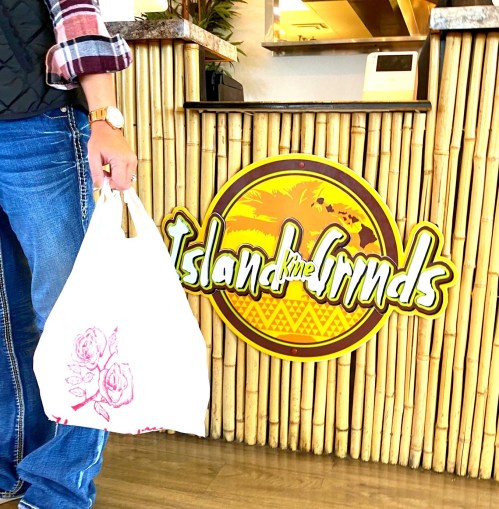 A person holding a takeout bag stands in front of a bamboo-themed restaurant sign that reads "Island Grinds."
