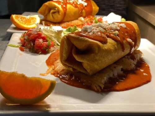 A plate of enchiladas topped with red sauce, cheese, and served with a side of salsa and orange slices.
