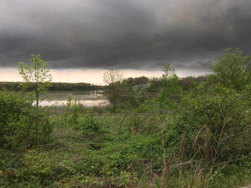 A serene landscape with a lake surrounded by greenery under a dramatic, cloudy sky.