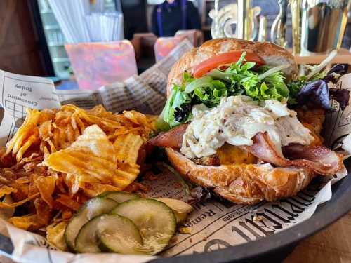 A croissant sandwich filled with lettuce, tomato, bacon, and chicken salad, served with a side of potato chips and pickles.