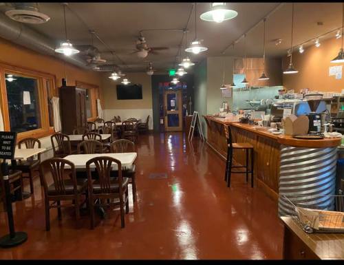A cozy café interior with wooden tables, chairs, a counter, and warm lighting. A sign requests customers to wait to be seated.