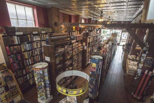 A cozy bookstore interior with tall wooden shelves filled with books, a spiral display, and large windows letting in light.
