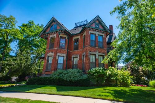 A charming red brick Victorian house surrounded by lush greenery and trees under a clear blue sky.