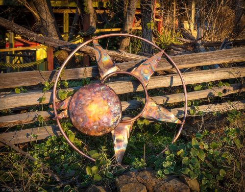 A decorative metal sun sculpture with colorful rays, set against a wooden fence and greenery.