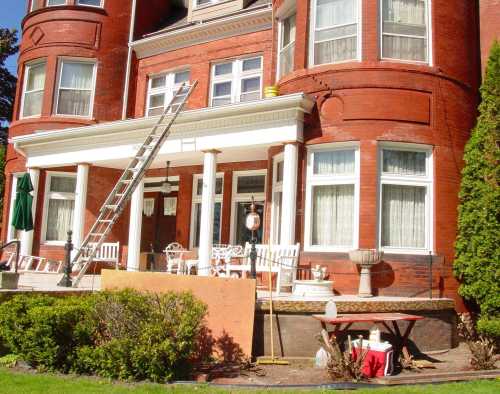 A red brick building with a ladder against it, porch furniture, and a garden area in front.