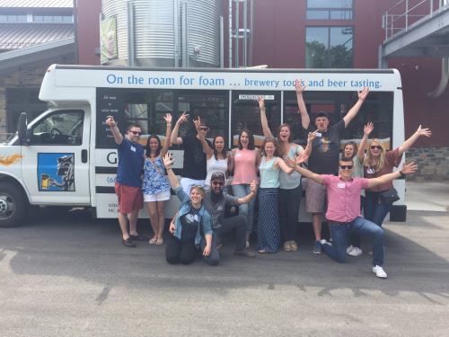 A group of people joyfully posing in front of a brewery tour bus, celebrating with raised arms and smiles.