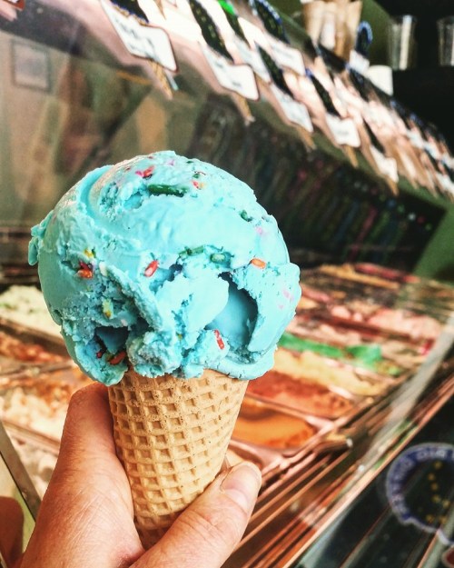 A hand holding a cone with bright blue ice cream, with various flavors displayed in the background.