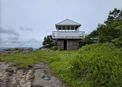 A stone fire lookout tower with a wooden deck, surrounded by greenery and rocky terrain under a cloudy sky.
