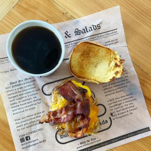 A cup of coffee beside a sandwich with bacon and cheese on a paper menu.