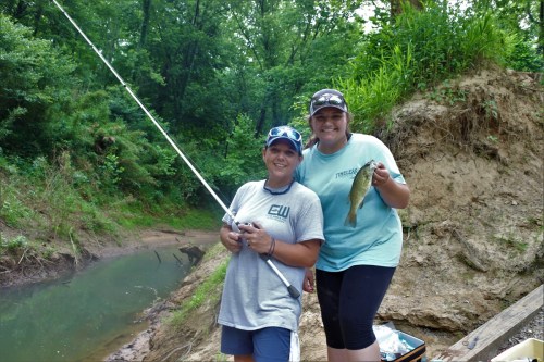 Two women stand by a creek, one holding a fishing rod and the other displaying a caught fish, surrounded by greenery.