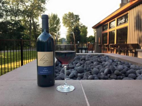 A bottle of 2014 Syrah wine next to a glass filled with red wine, set on a stone table outside a modern building.