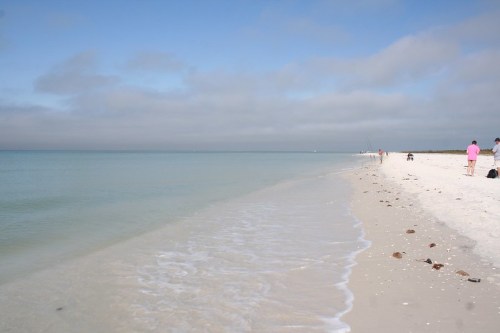 A serene beach scene with soft waves, white sand, and a cloudy sky, featuring a few distant beachgoers.