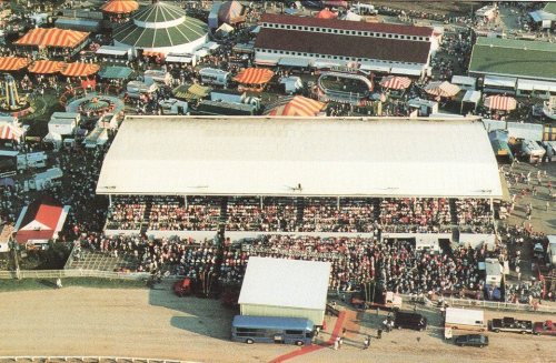 Aerial view of a crowded fairground with colorful tents, rides, and a large audience gathered in front of a grandstand.