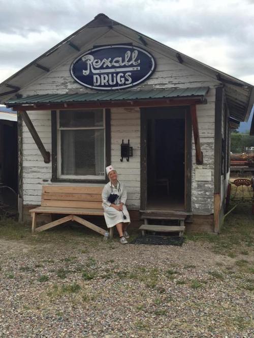A person in a white coat sits on a bench outside an old Rexall Drugs store, surrounded by gravel and rustic scenery.