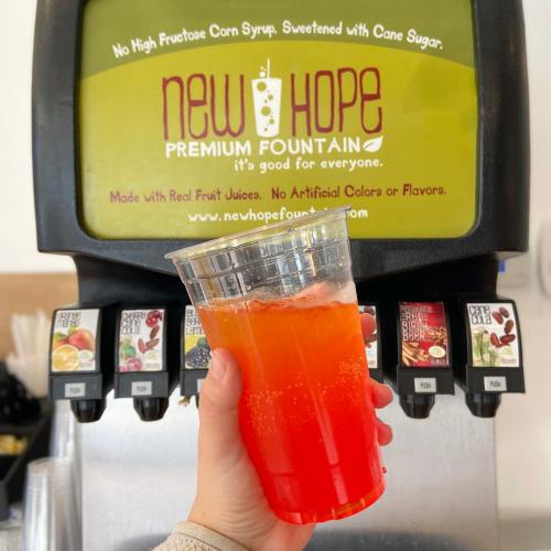 A hand holds a bright red drink in front of a beverage dispenser with a sign for New Hope Premium Fountain.