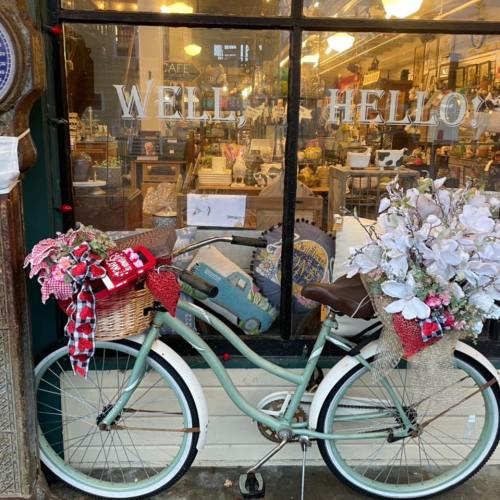 A vintage bicycle with a basket, decorated with flowers, parked in front of a charming shop window that says "WELL HELLO."