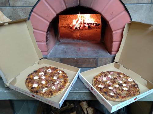 Two pizzas in open boxes sit in front of a wood-fired oven, with flames visible in the background.