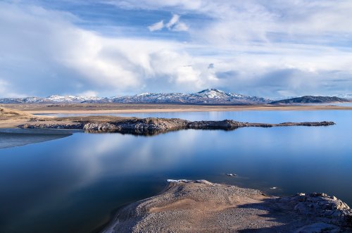 A serene landscape featuring a calm lake, rocky shores, and distant snow-capped mountains under a blue sky.