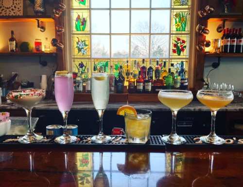 A colorful array of cocktails lined up on a bar, with a stained glass window in the background.