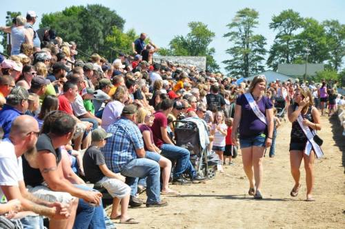 A large crowd watches two young women in sashes walking along a dirt path at an outdoor event on a sunny day.