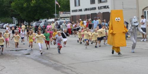 A lively street scene with children in yellow shirts running, accompanied by a large yellow mascot and a gray character.