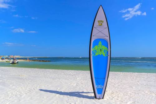 A colorful surfboard stands on a sandy beach with calm blue waters and a clear sky in the background.