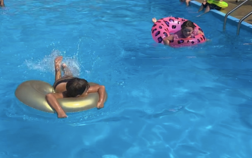Two children play in a pool, one on a gold float and the other on a pink float with a pattern, splashing water.