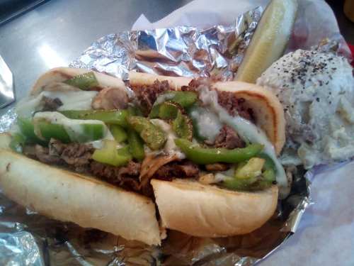A loaded sandwich with beef, green peppers, and melted cheese, served with a pickle and a side of potato salad.