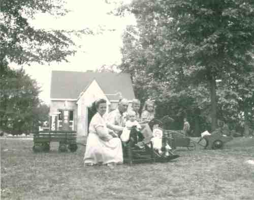 A black-and-white photo of a family sitting outdoors with children and wagons in front of a house surrounded by trees.