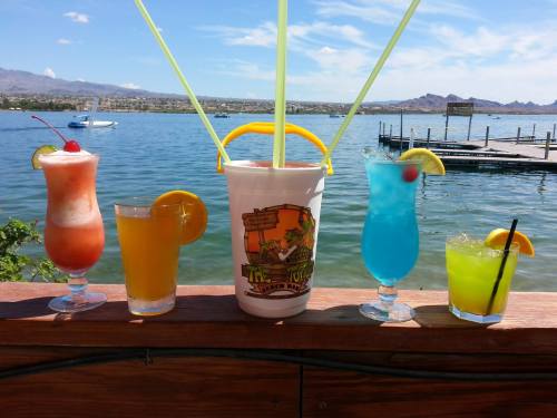 Five colorful cocktails in glasses, with a bucket of drinks in the center, set against a scenic lake view.