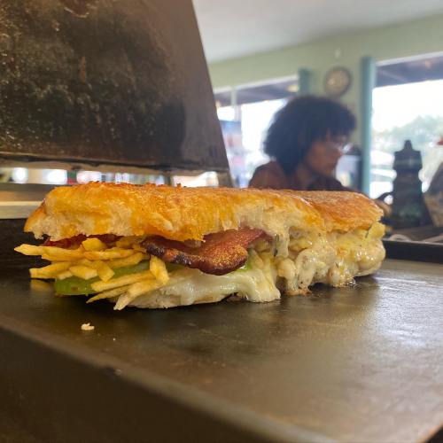 A grilled sandwich with bacon, cheese, and vegetables, resting on a hot griddle in a cozy café setting.
