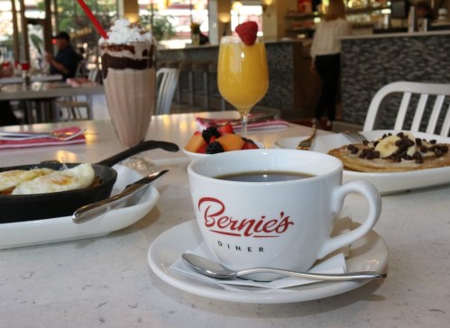 A cozy diner table with coffee, a fruit cup, a milkshake, and a breakfast skillet, featuring "Bernie's Diner" mug.