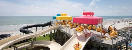 A water park with colorful slides and visitors enjoying the beach in the background under a clear blue sky.