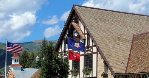 A building with a steep roof, featuring flags, surrounded by trees and mountains under a blue sky.
