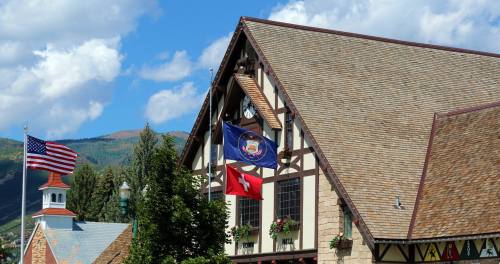 A charming building with a steep roof, featuring flags and surrounded by trees against a blue sky with clouds.